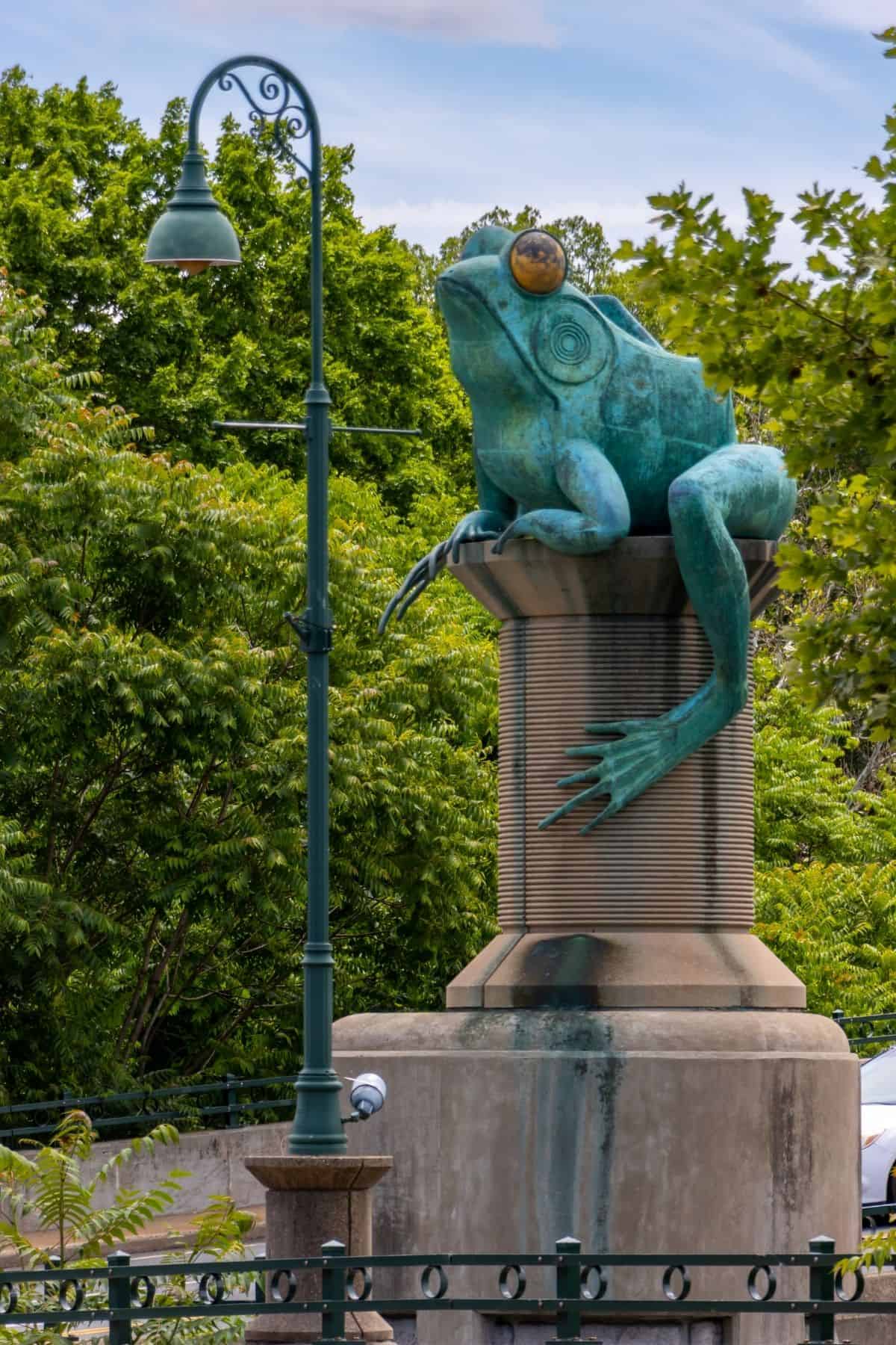 A large frog statue sits atop a pedestal next to a green streetlamp, surrounded by trees and greenery.