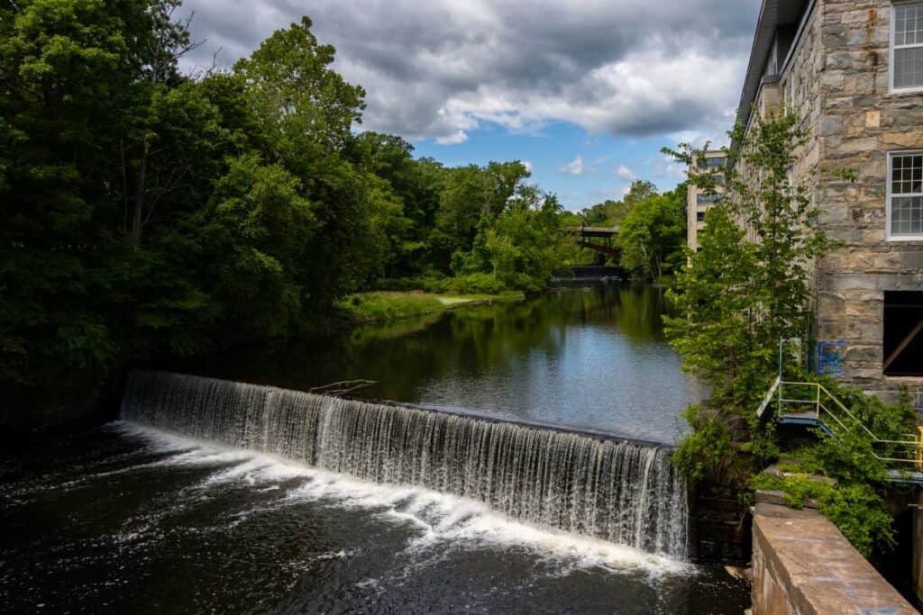 A small waterfall flows over a low dam in a river, surrounded by green trees and a stone building on the right under a partly cloudy sky.