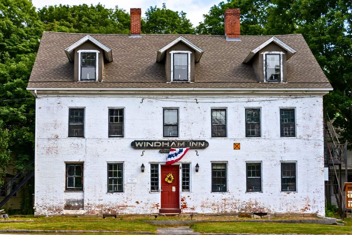 A white, two-story brick building labeled “Windham Inn” with a brown shingle roof, six chimneys, and a decorated front door, standing in front of green trees.