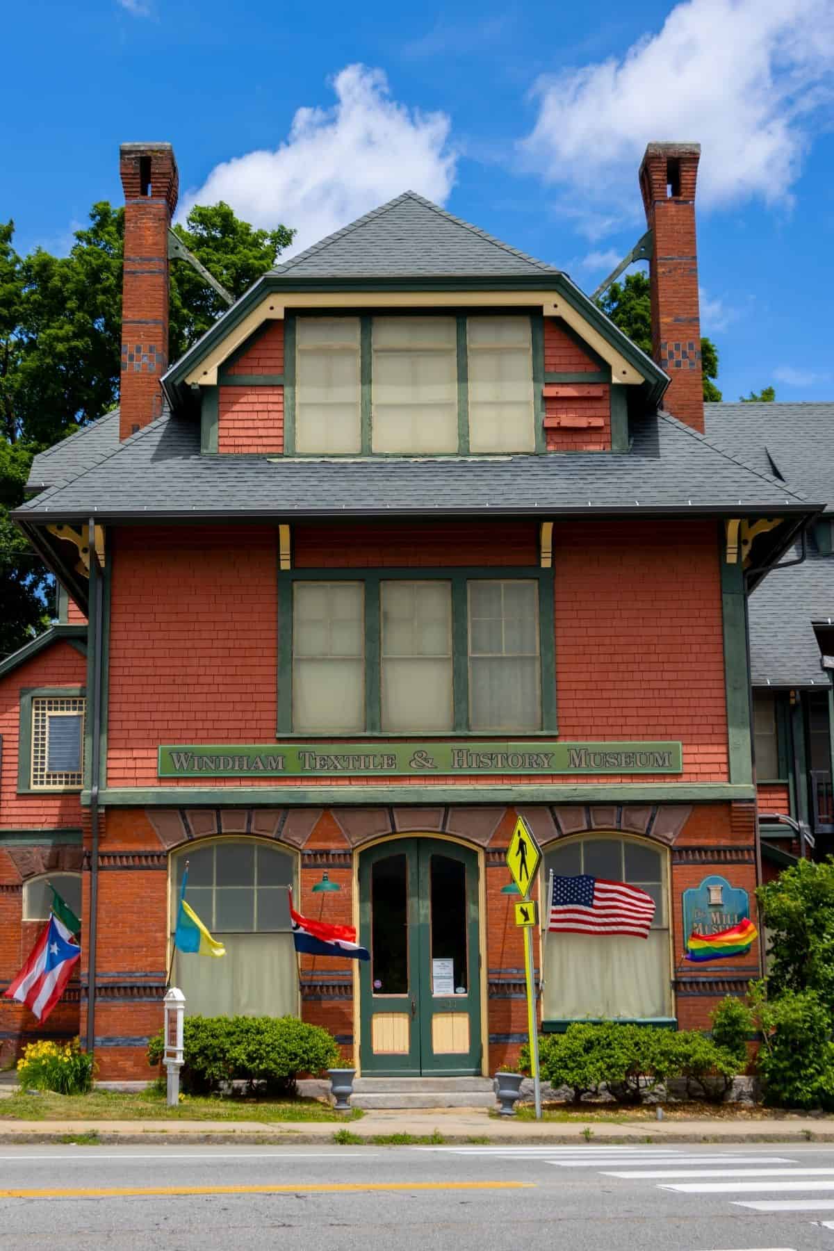 A red brick building labeled “Windham Textile & History Museum” displays several flags outside and sits along a street with a pedestrian crossing sign.