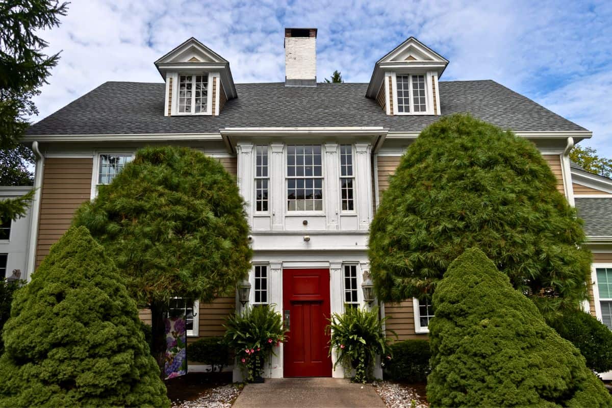 Two-story house with tan siding, white trim, a red front door, two dormer windows, and large trimmed bushes in front, under a partly cloudy sky.