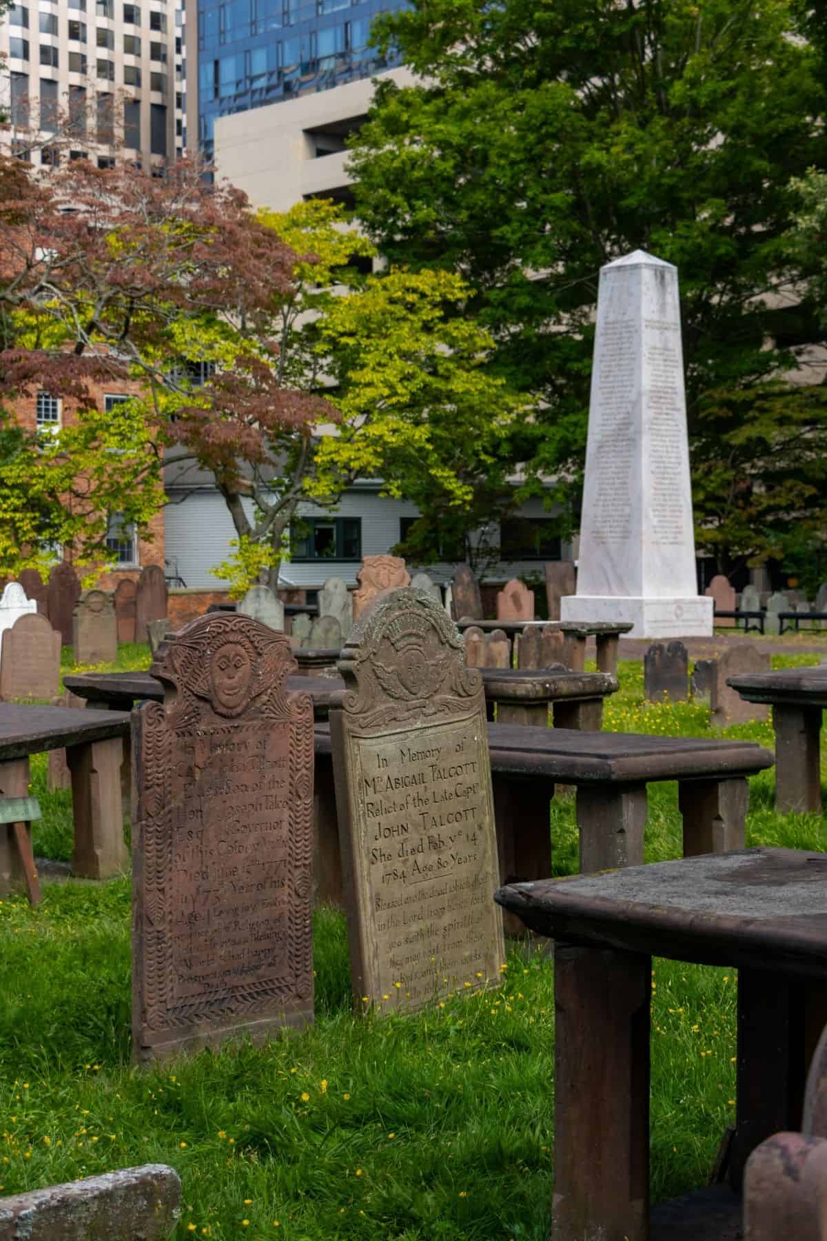 Old gravestones and an obelisk memorial stand in a historic cemetery with green grass, benches, and city buildings visible in the background.