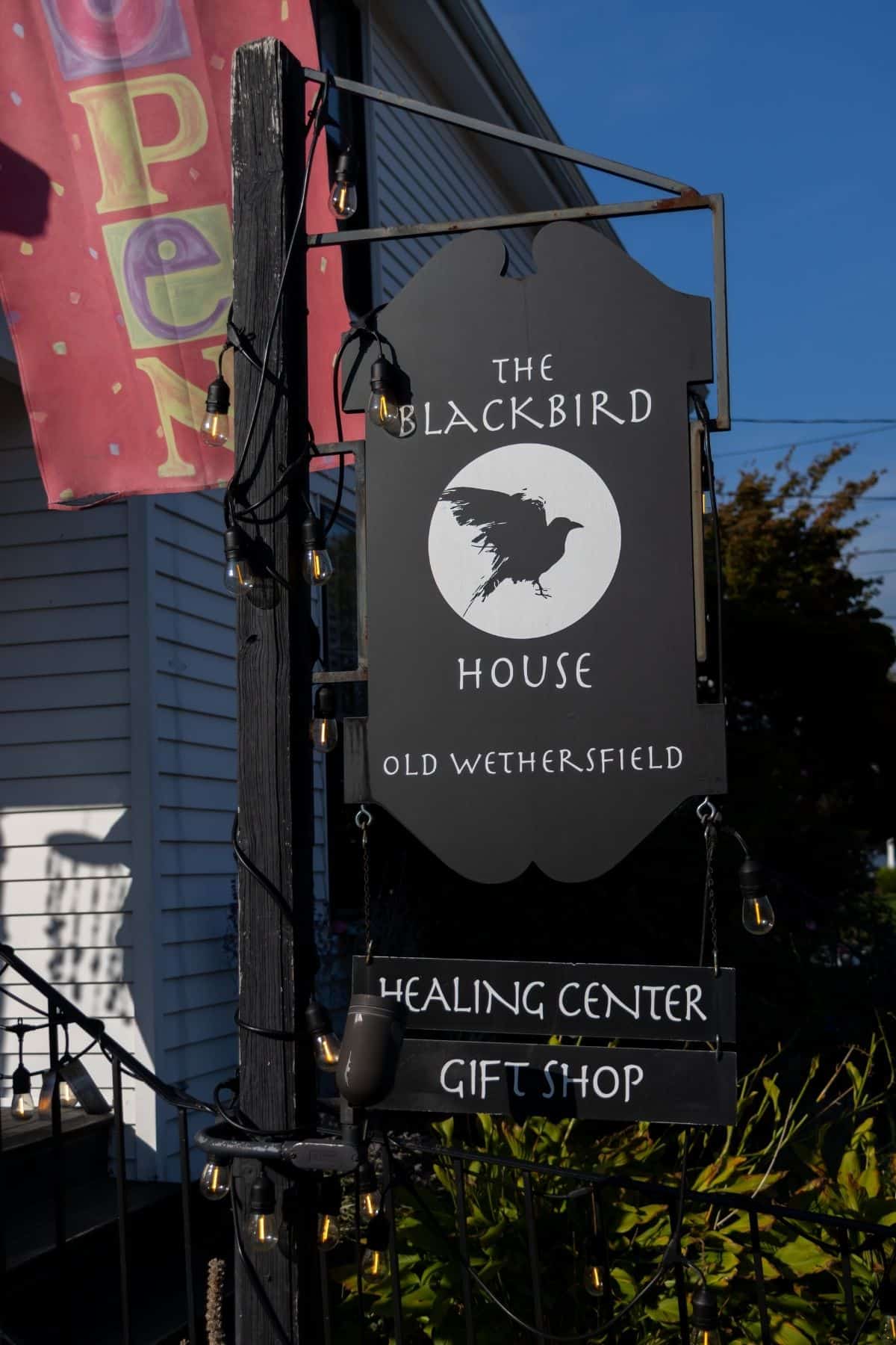 A sign for The Blackbird House in Old Wethersfield reads “Healing Center, Gift Shop” with a blackbird silhouette. Outdoor string lights decorate the signpost.