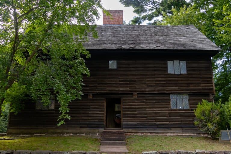A two-story, dark wooden colonial house with diamond-paned windows, a central chimney, and trees partially shading the front façade.