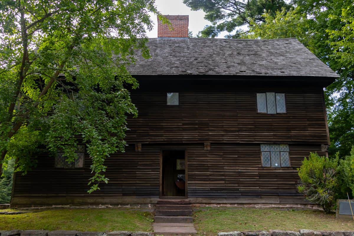 A two-story, dark wooden colonial house with diamond-paned windows, a central chimney, and trees partially shading the front façade.