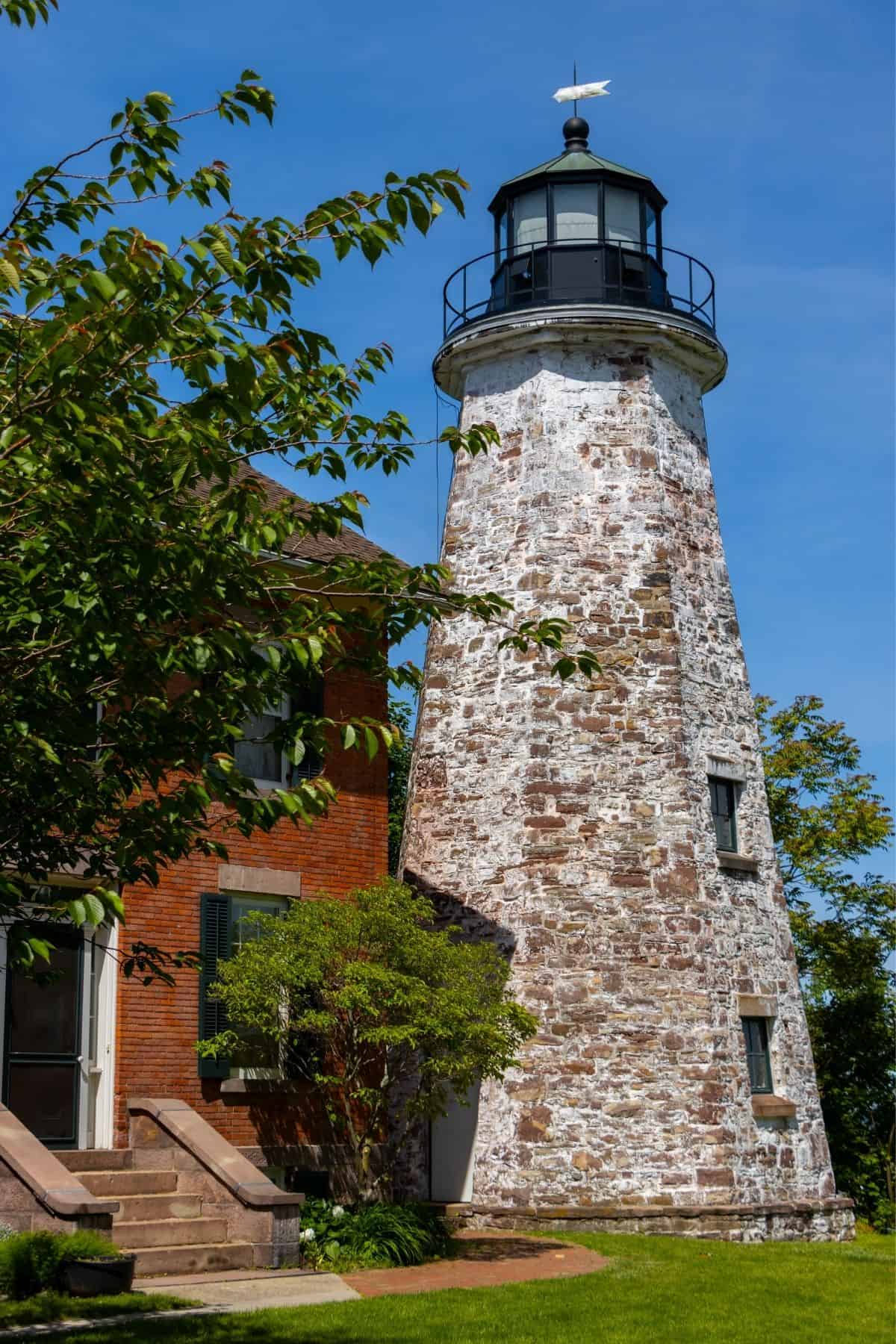 A tall, weathered stone lighthouse stands beside a brick building, surrounded by green grass and trees under a clear blue sky.
