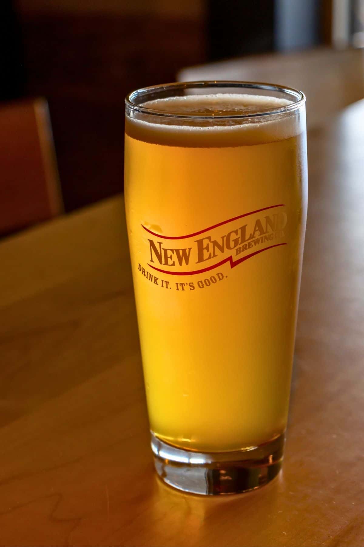 A pint glass filled with golden beer sits on a wooden table. The glass is branded with “New England Brewing” and the slogan “Drink it. It’s good.”.