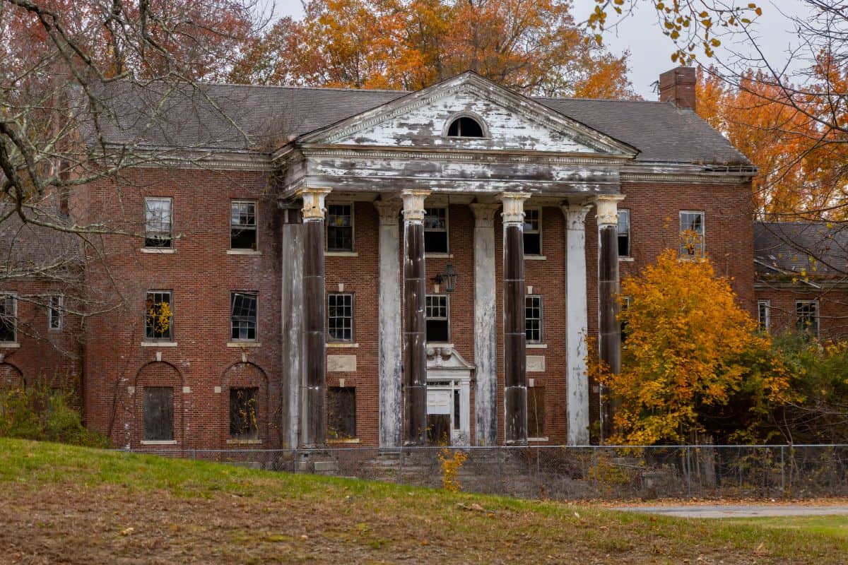 An abandoned brick building with tall columns and boarded windows, surrounded by autumn trees with orange and yellow leaves.