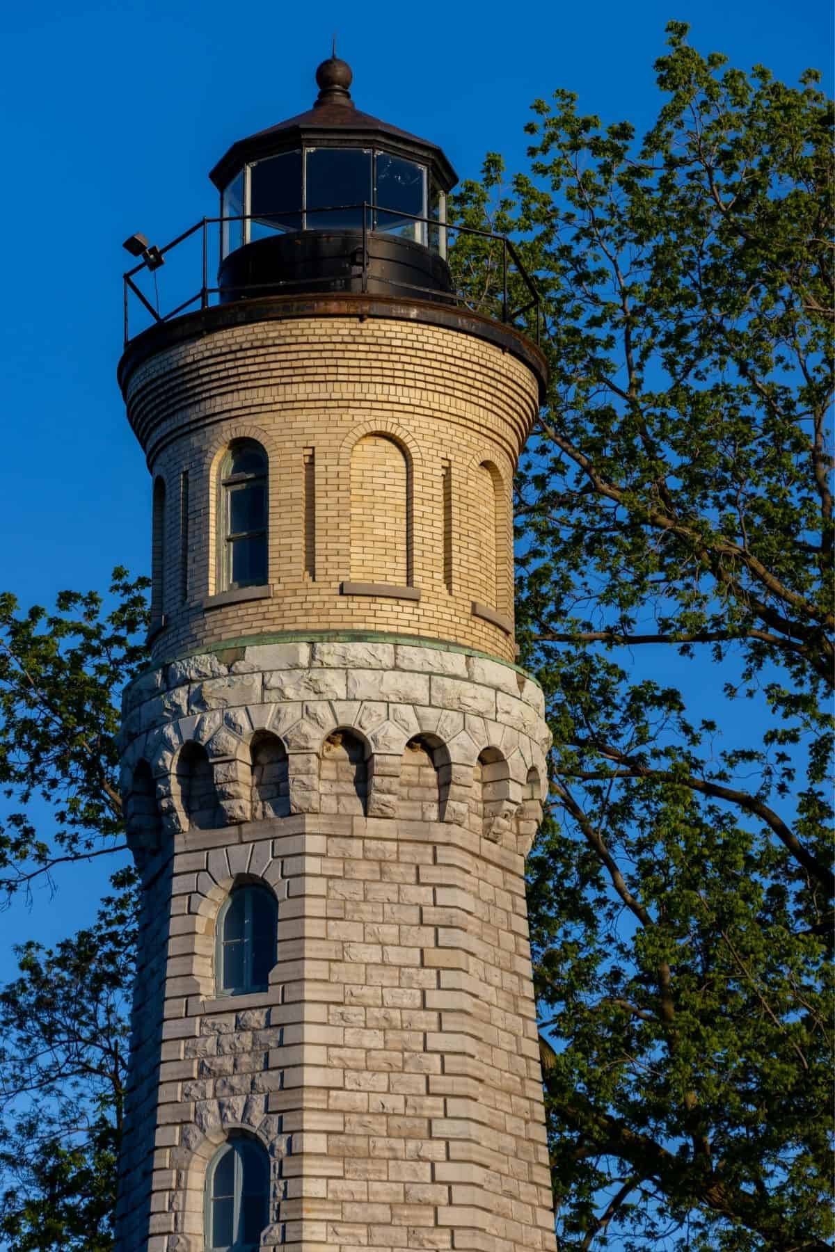 Tall stone lighthouse with a cylindrical tower and black lantern room, set against a clear blue sky and partially framed by leafy green tree branches.