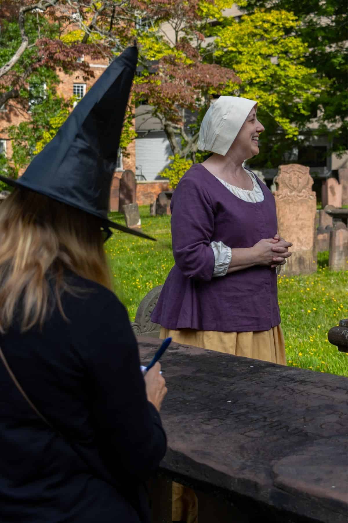 A woman in historical clothing stands and smiles in a graveyard, while another person in a black witch hat sits in the foreground, writing on a notepad.