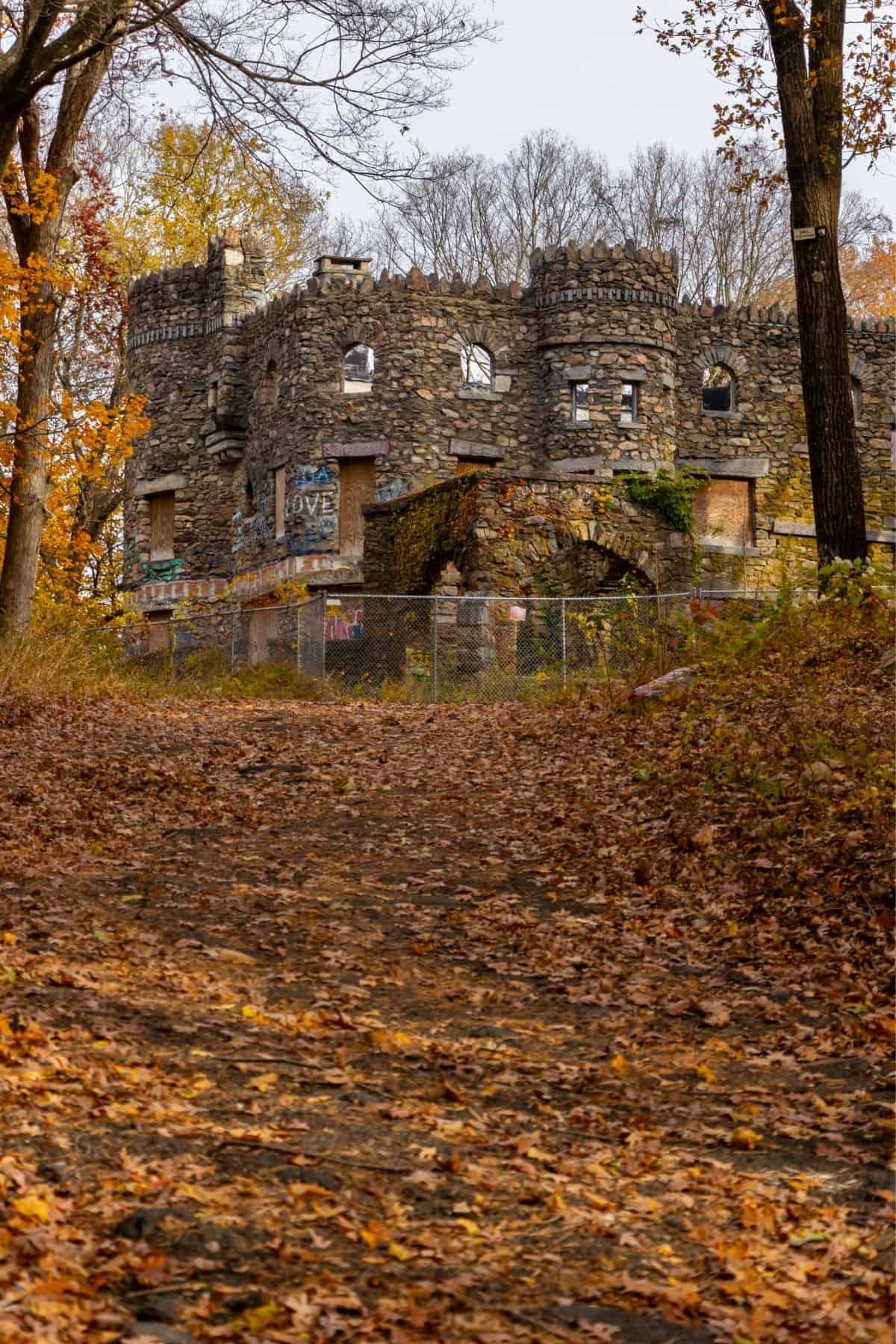 A stone castle ruin with boarded windows and graffiti stands in a forest with autumn leaves covering the ground and trees without many leaves.