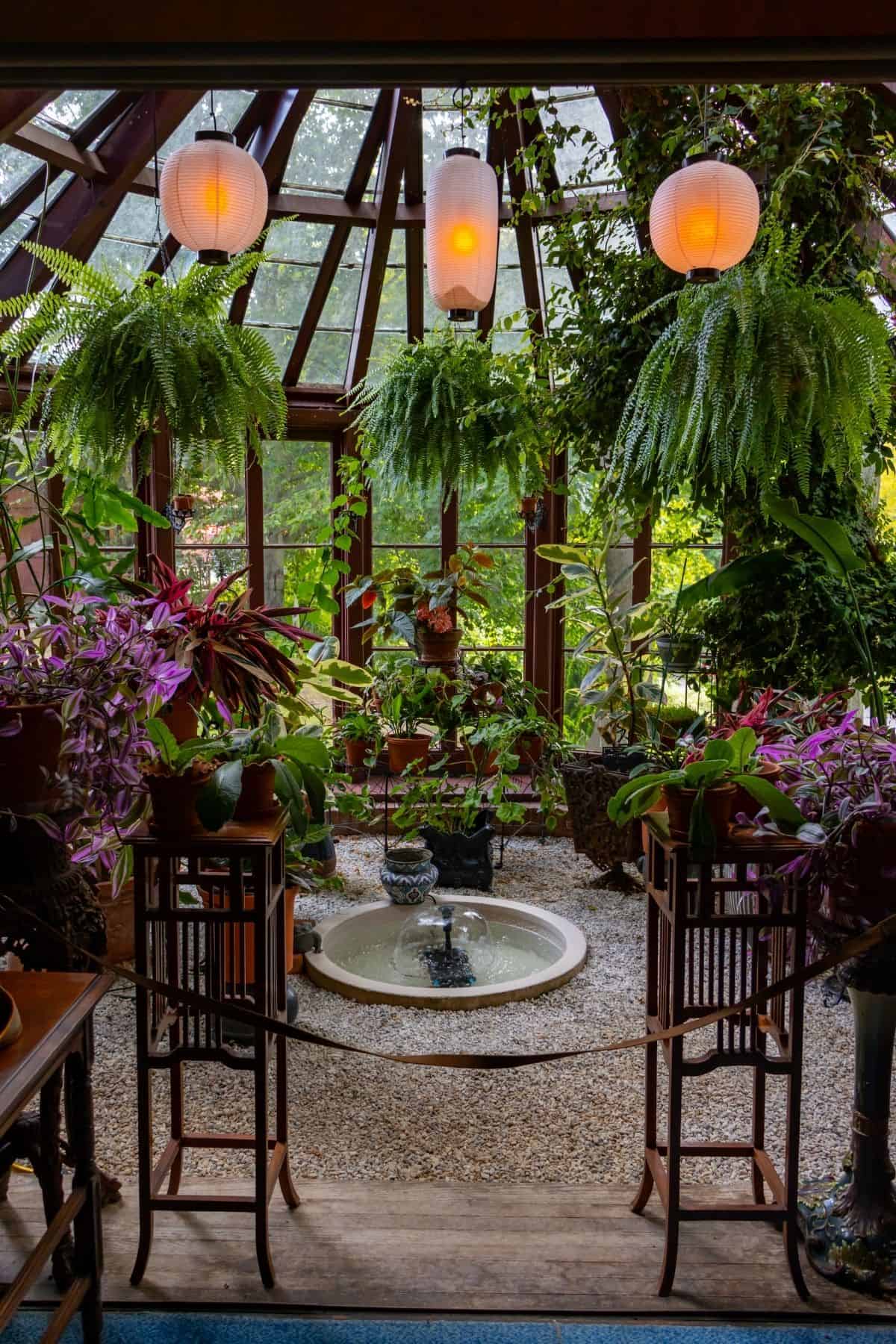 Indoor garden with hanging ferns, assorted potted plants, a small central fountain, wooden chairs, and three hanging lanterns under a glass ceiling.