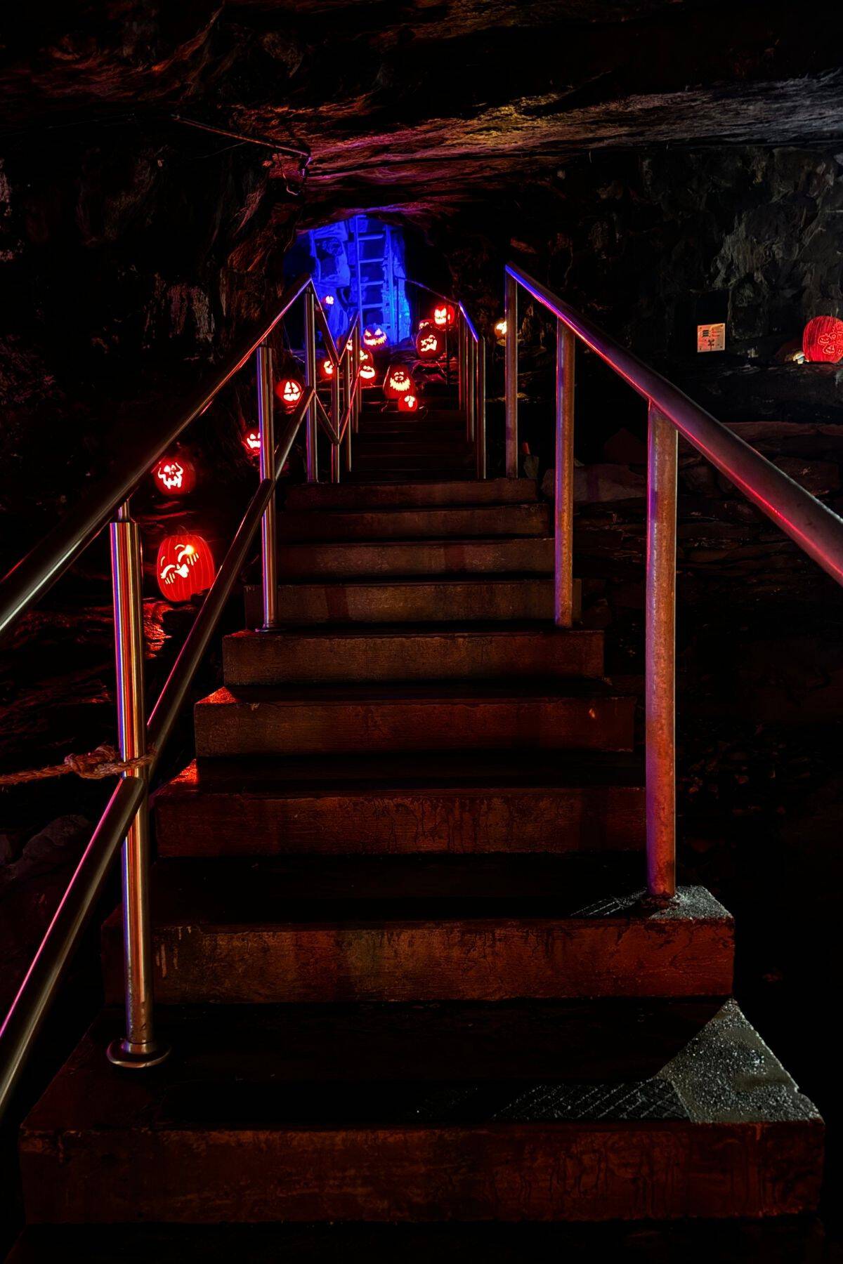 A dimly lit stairway with metal railings leads upward, flanked by glowing carved pumpkins. The tunnel walls are dark, and blue light illuminates the background.