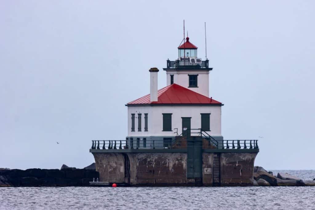 A white lighthouse with a red roof stands on a concrete platform surrounded by water, with rocks and overcast sky in the background.
