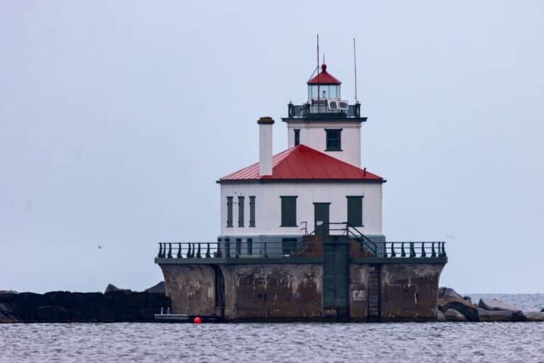 A white lighthouse with a red roof stands on a concrete platform surrounded by water, with rocks and overcast sky in the background.