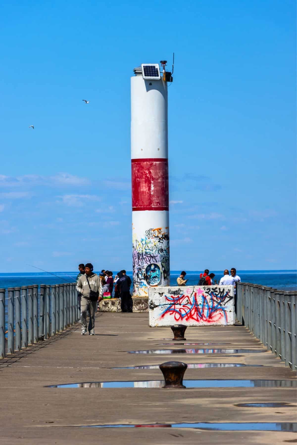 People walk and fish on a pier with puddles, leading to a red and white striped lighthouse covered in graffiti, under a clear blue sky.