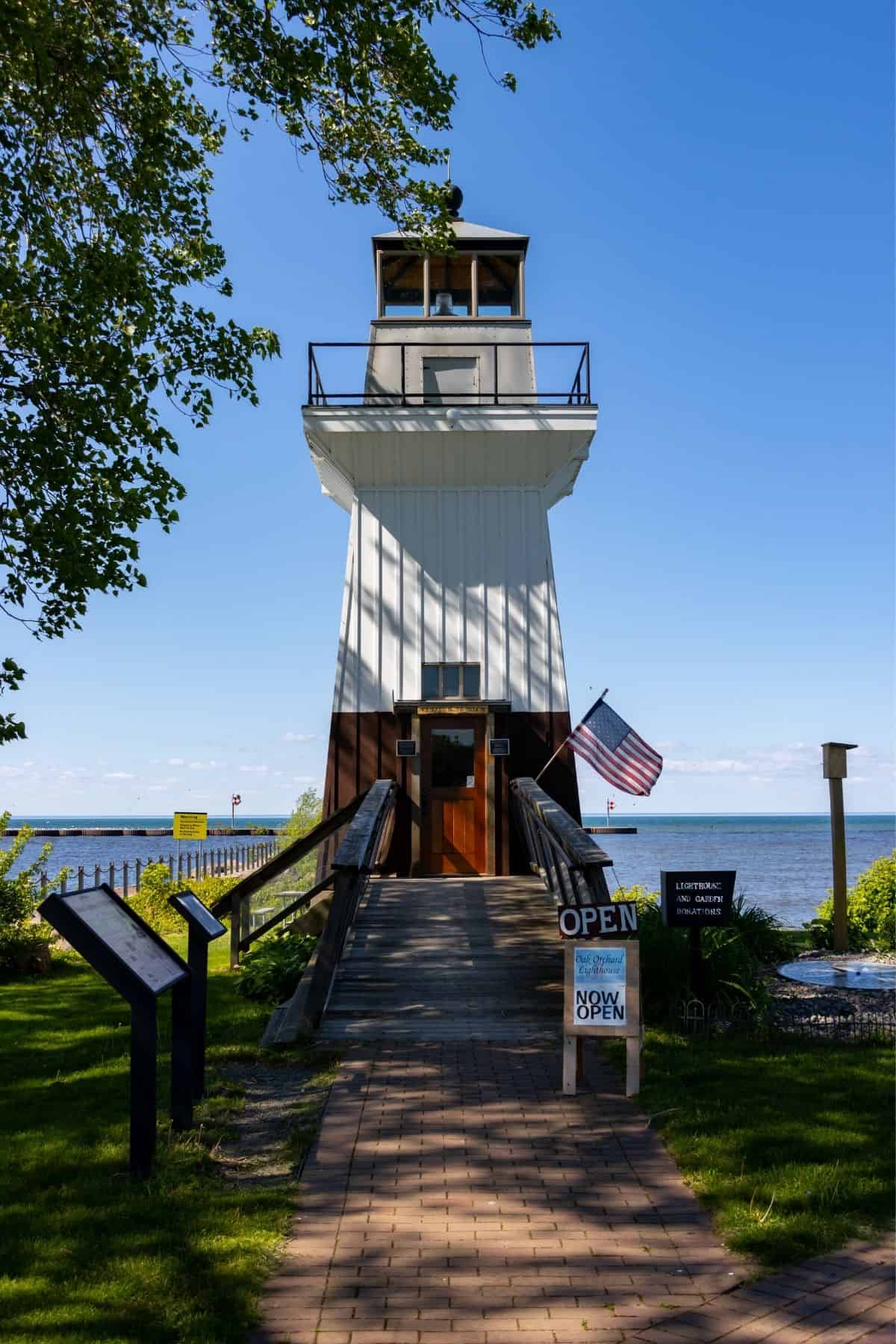 A white lighthouse with a brown base stands by the water, surrounded by greenery. An American flag and “OPEN” signs are visible near the entrance.