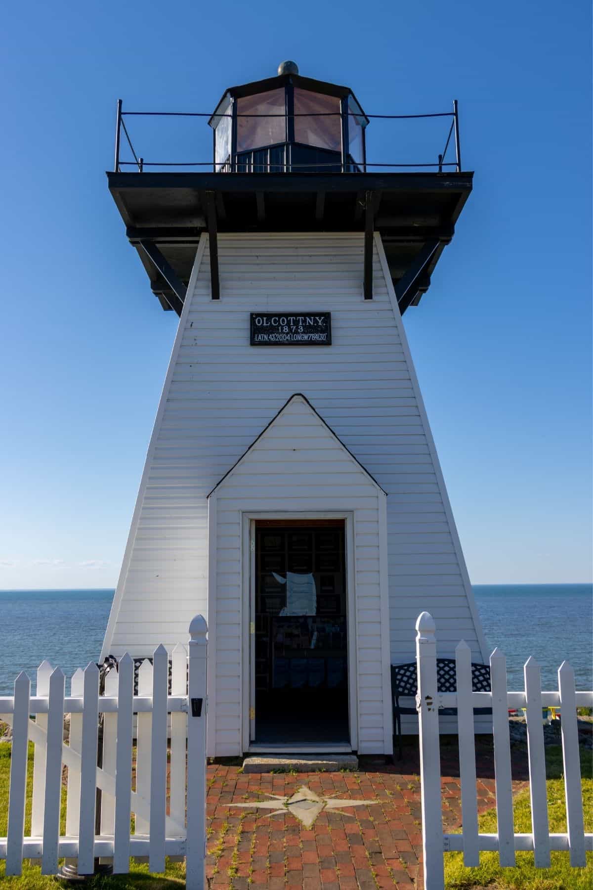 A white wooden lighthouse with a black lantern room stands by the ocean, viewed through an open white picket fence gate under a clear blue sky.