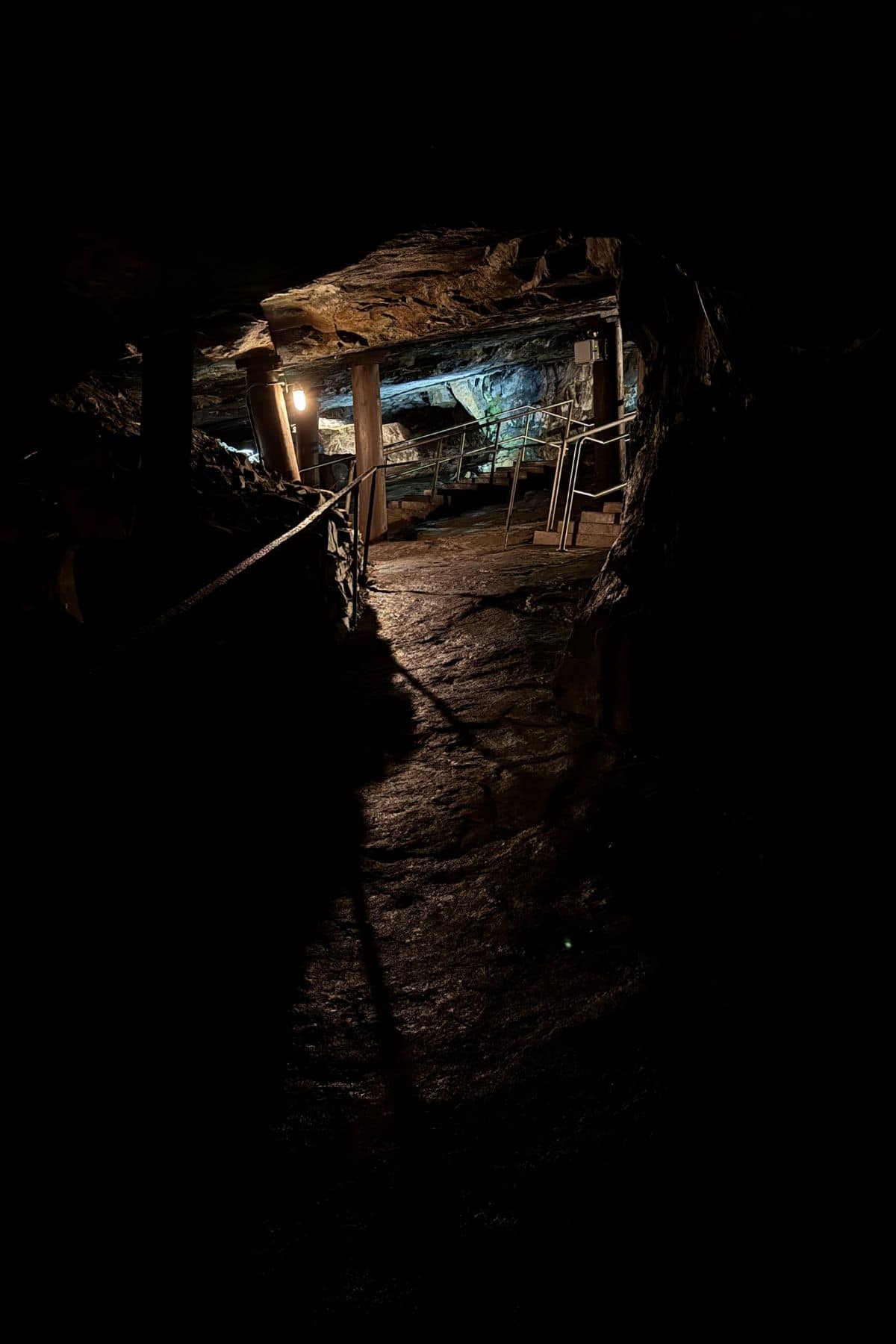 Dimly lit underground tunnel with rocky walls, wooden supports, handrails, and artificial lighting visible further in the passage.