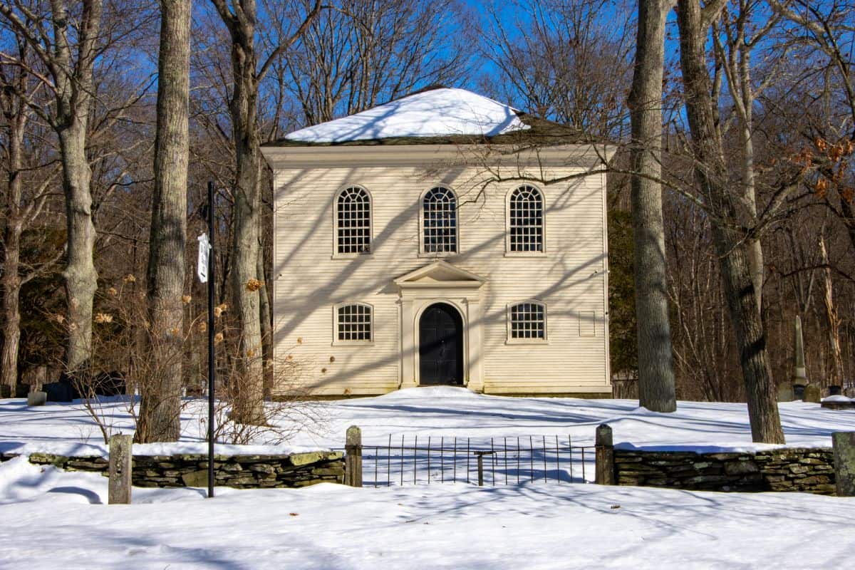 A small, beige, two-story building with arched windows stands among leafless trees and snow-covered ground under a clear blue sky.