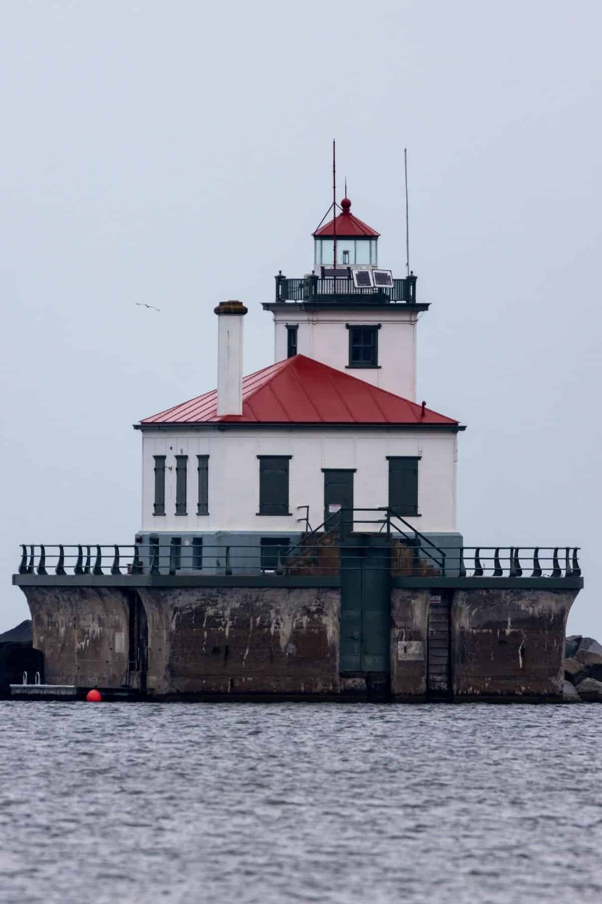 White lighthouse with a red roof sits on a concrete platform surrounded by water, under an overcast sky.