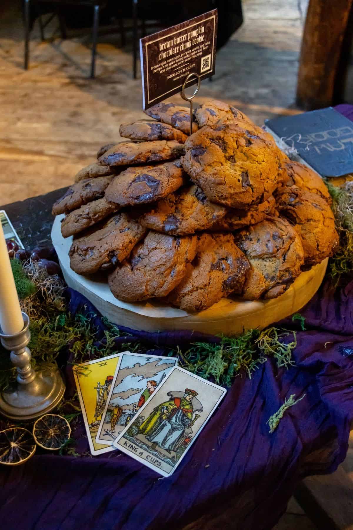 A pile of chocolate chip cookies on a tray, labeled with a sign, sits on a purple cloth near three tarot cards and a candle holder.