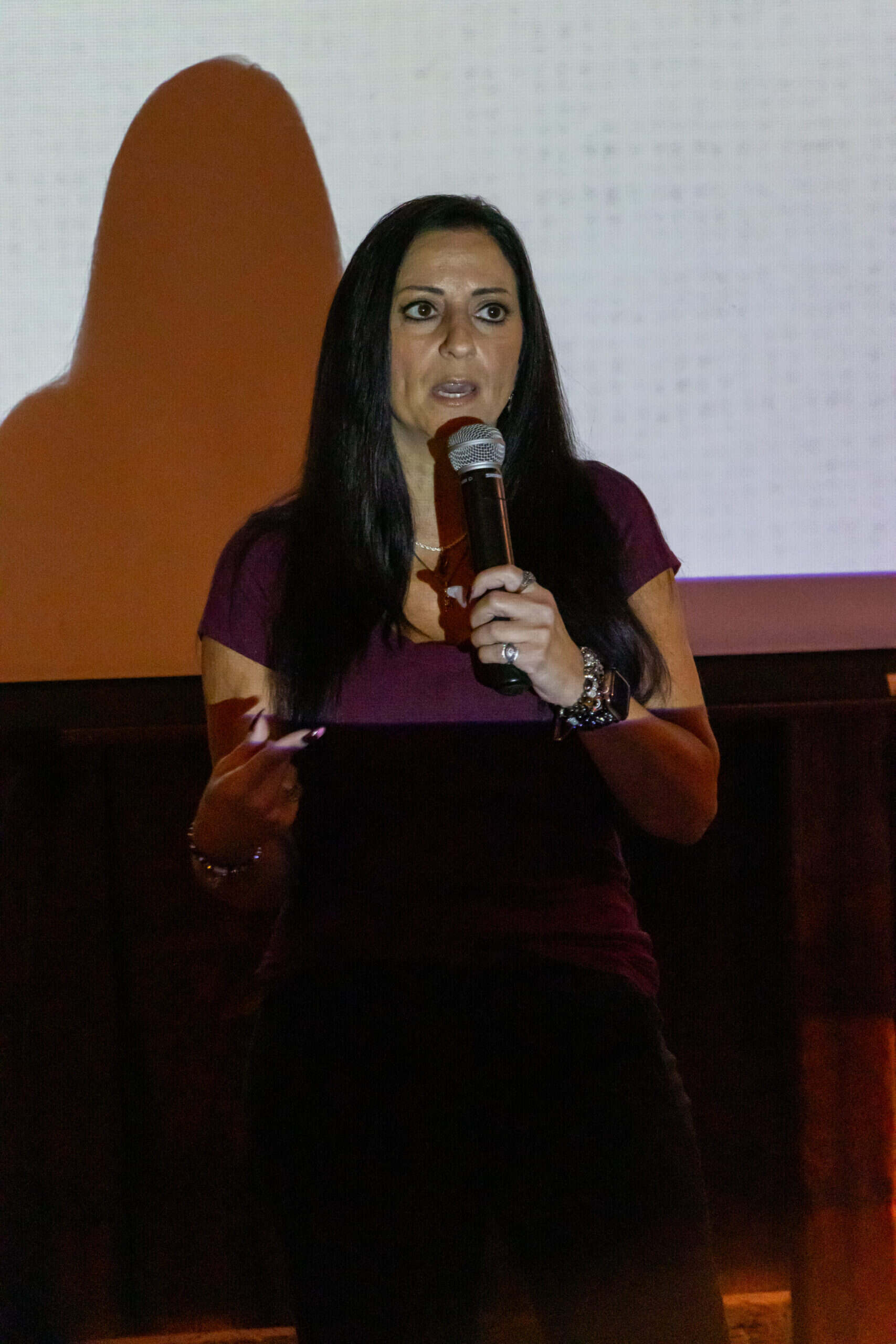 A woman with long dark hair speaks into a microphone while standing in front of a projection screen.