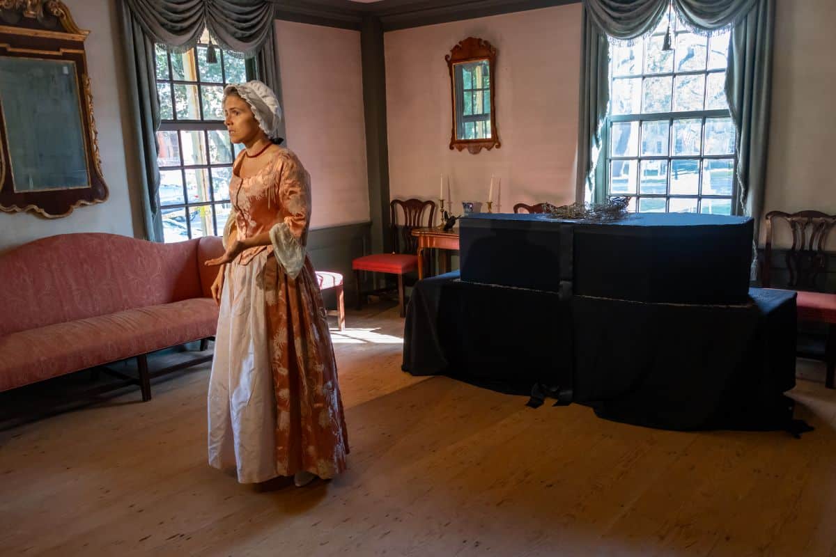A woman in colonial-era dress stands in a historic room with antique furniture, wooden floor, large windows, and a covered object in the corner.