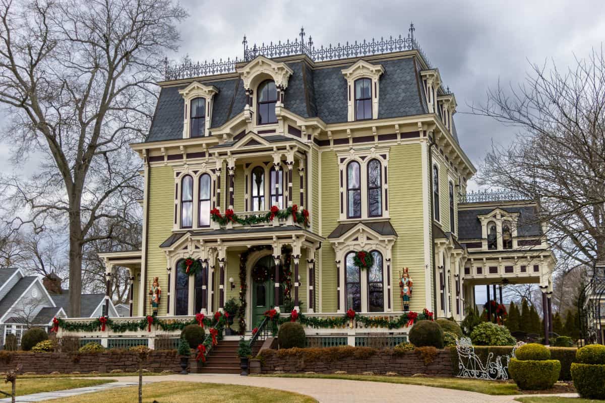 Large Victorian-style house with ornate trim, decorated with red and green holiday wreaths and garlands, set against a cloudy sky and surrounded by leafless trees and manicured bushes.