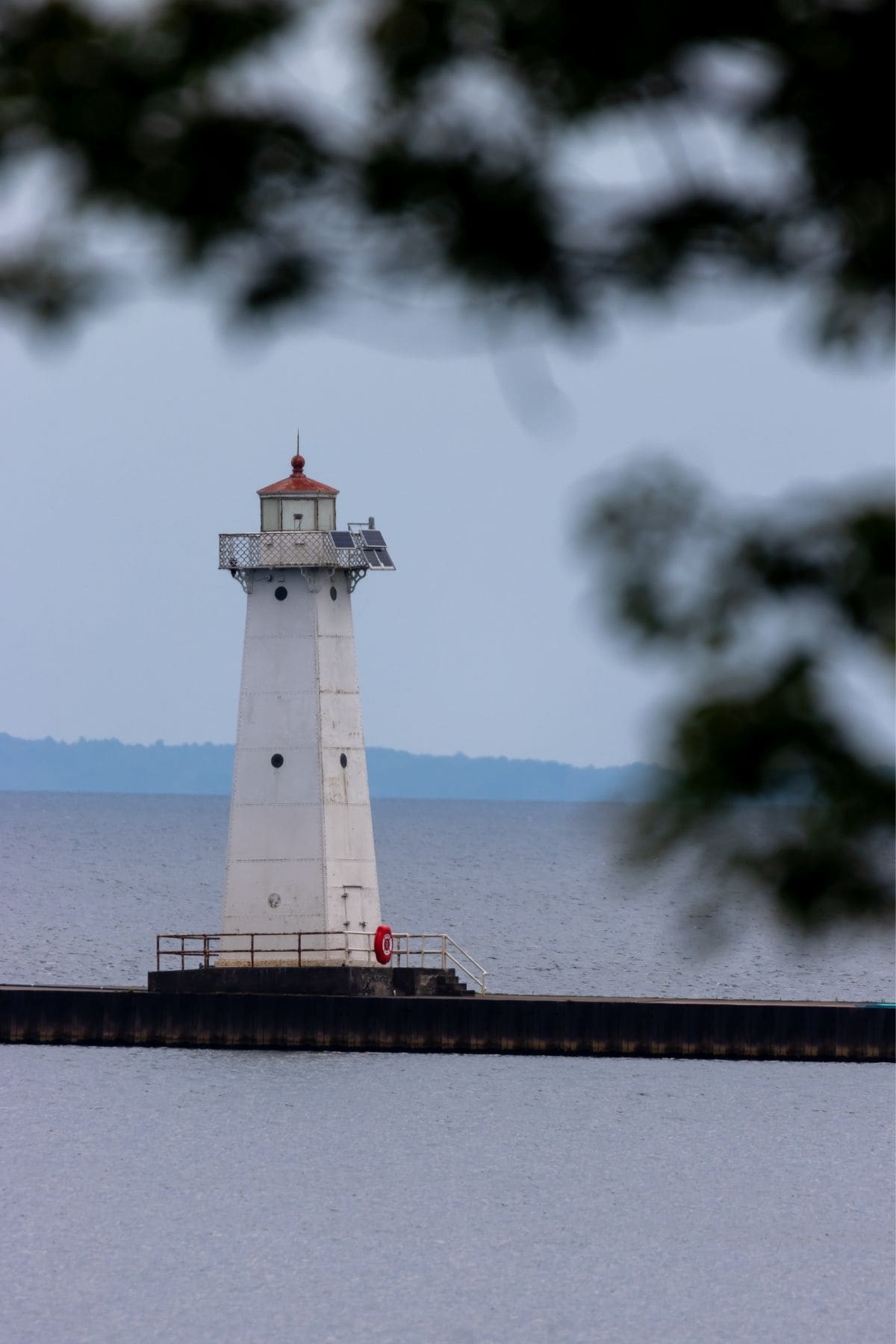 A white lighthouse with a red roof stands on a pier by calm water, with distant land visible in the background and tree branches framing the view.