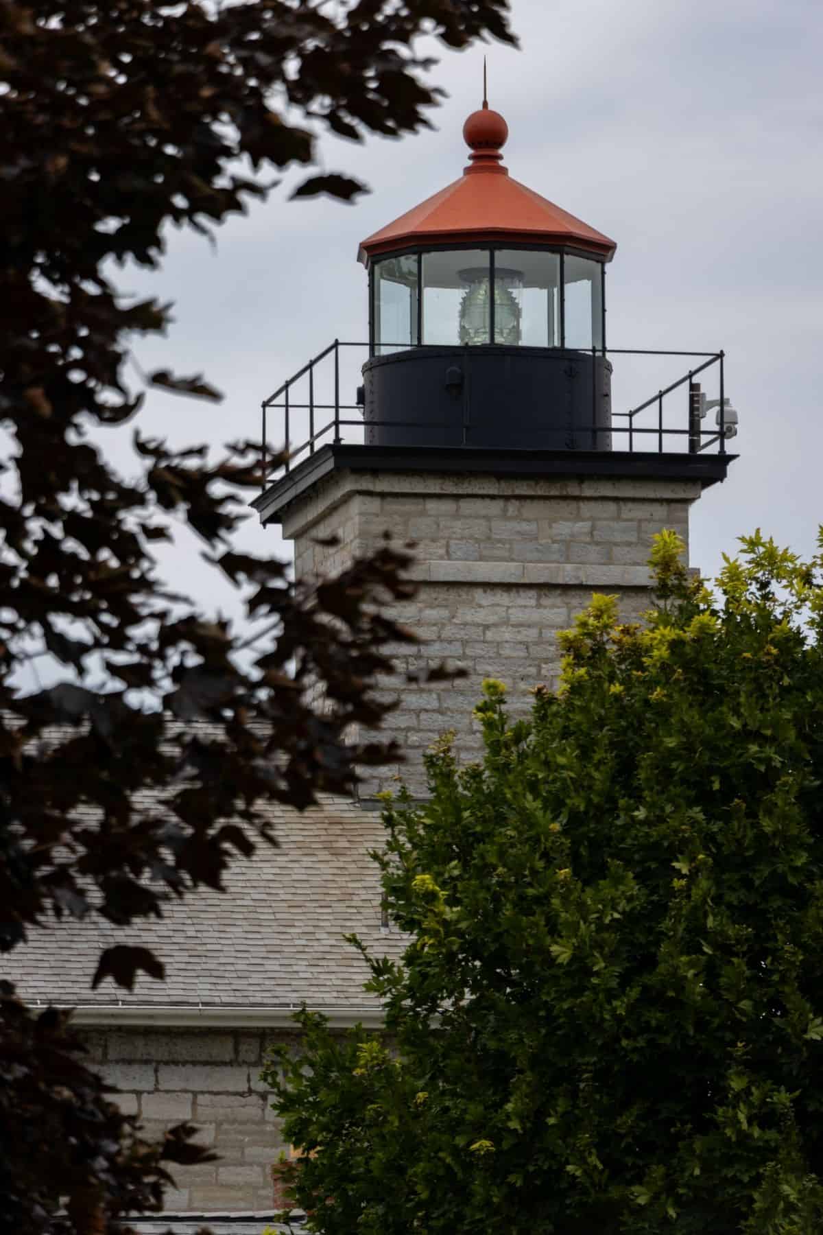 A stone lighthouse with a red roof seen partially behind leafy green trees, under a cloudy sky.