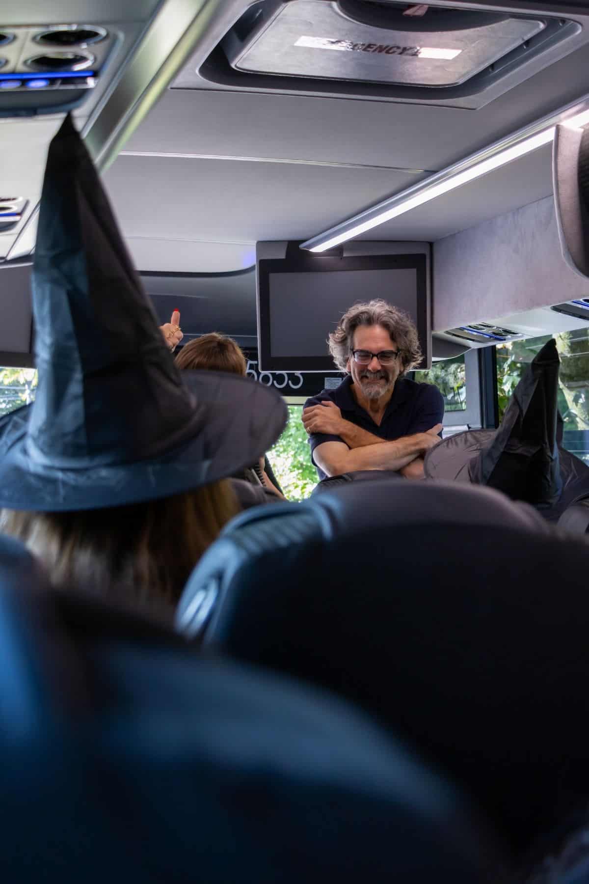 A person in a witch hat sits on a bus, facing a man with glasses and gray hair who is leaning on a seat and smiling.