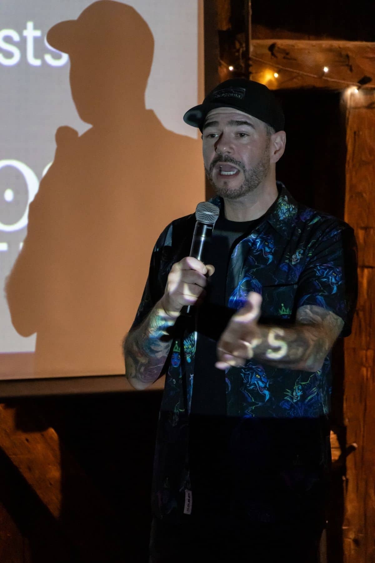 A man in a patterned shirt and cap holds a microphone and gestures while speaking on stage, with his shadow visible on a screen behind him.