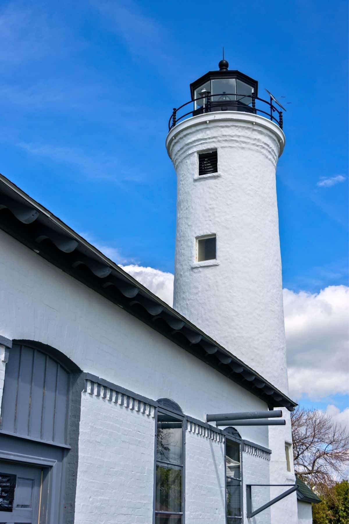White lighthouse tower with black lantern rises beside a white brick building under a blue sky with clouds.
