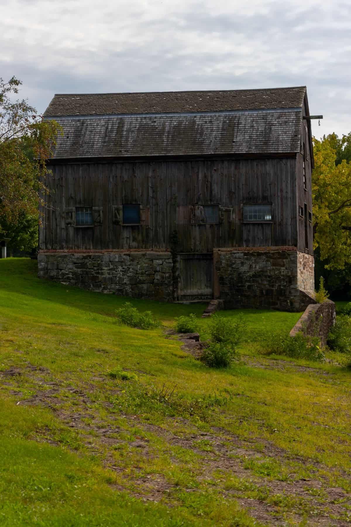 A large, weathered wooden barn with small windows sits atop a stone foundation, surrounded by green grass and trees under a cloudy sky.
