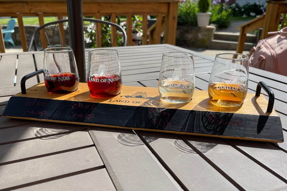 A wooden tray holds four labeled tasting glasses with different colored drinks, set on an outdoor table in sunlight.