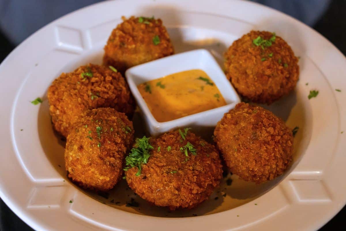 Five breaded, fried appetizer balls arranged around a small central bowl of dipping sauce in a white bowl.