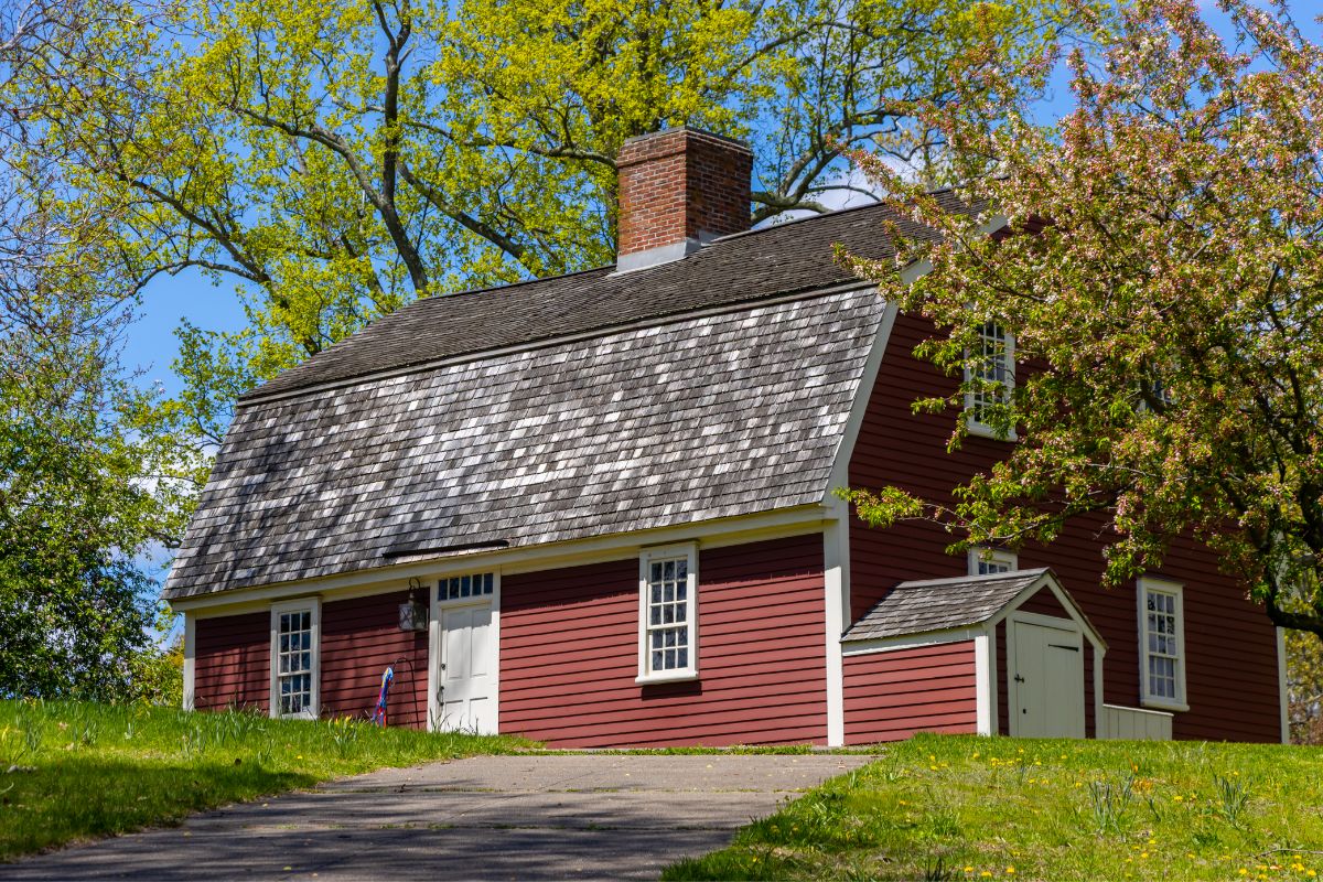 A red historic house with a gambrel roof, set on a grassy hill surrounded by trees.