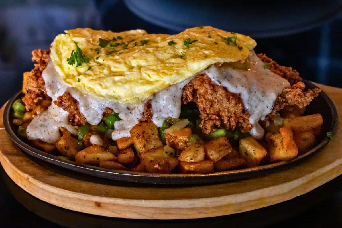 Fried chicken topped with an omelet and gravy over a bed of roasted potatoes and peas on a metal skillet.
