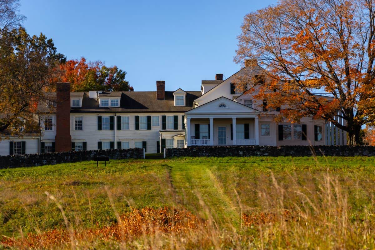 A large white farmhouse with a stone wall and fall trees, viewed across a grassy field.