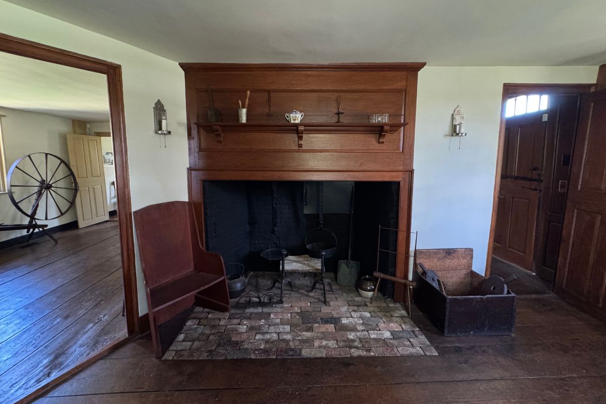A brick fireplace with a wooden mantel, flanked by a wooden chair, a spinning wheel in the doorway, and a wooden chest.