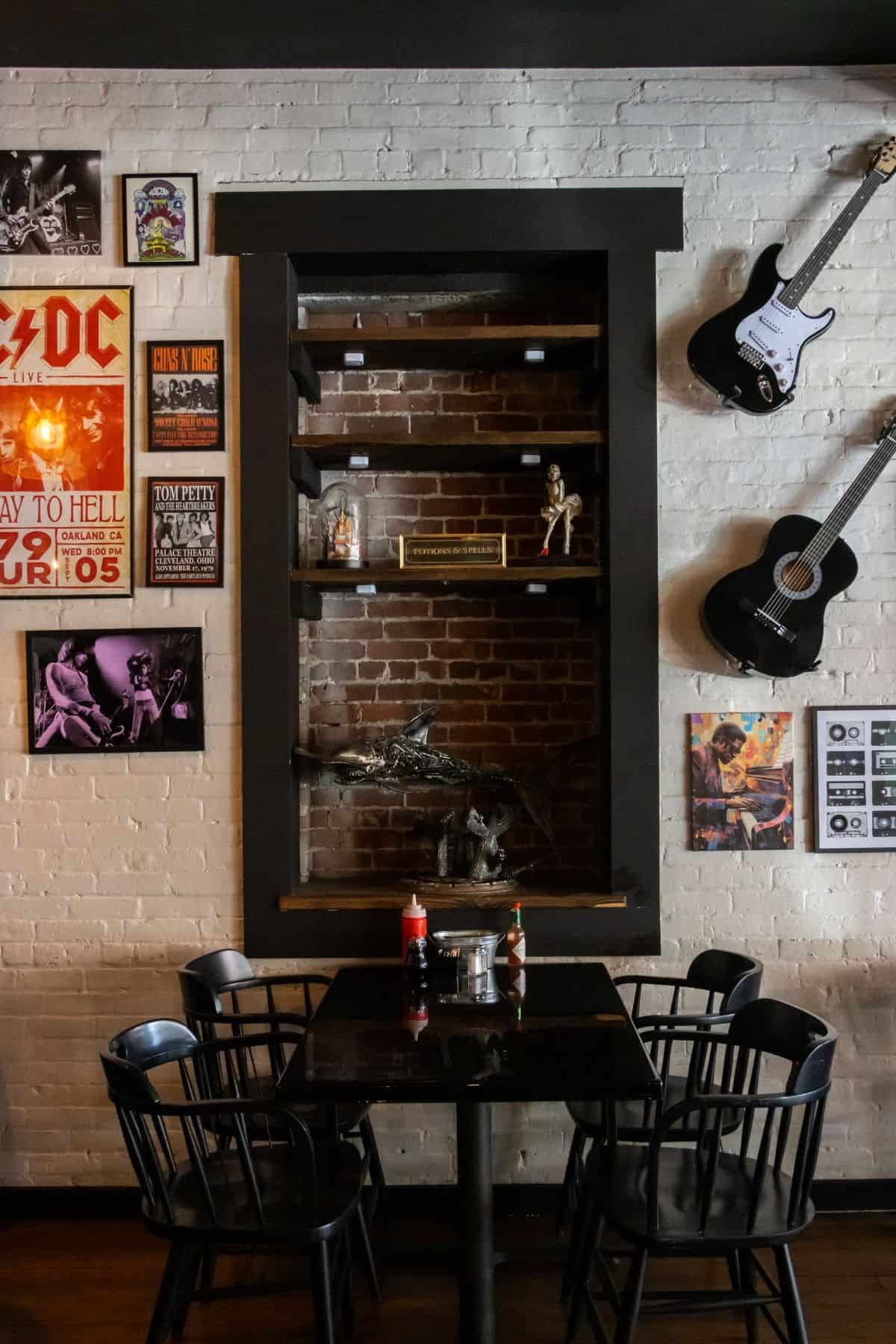 A square dining table with four black chairs placed against a brick wall decorated with music posters, two guitars, and a central built-in shelf containing small decorative items.