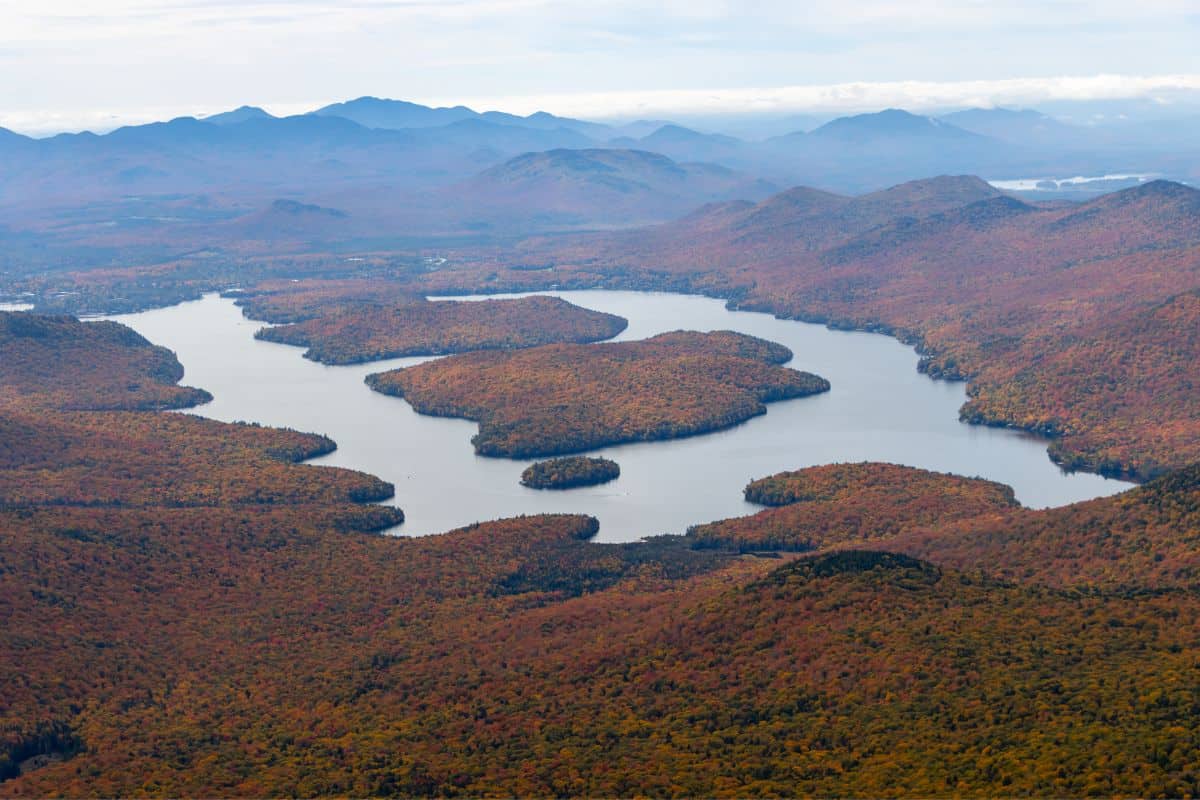 Aerial view of a lake with islands surrounded by autumn-colored forest and mountain ranges in the distance.
