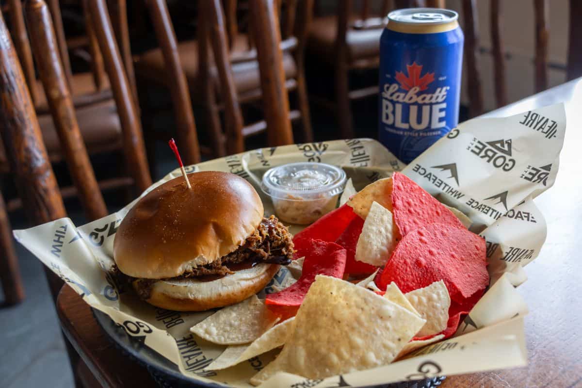 A pulled pork sandwich with a red toothpick, a side of tortilla chips, and a small container of dip, served with a can of Labatt Blue beer.