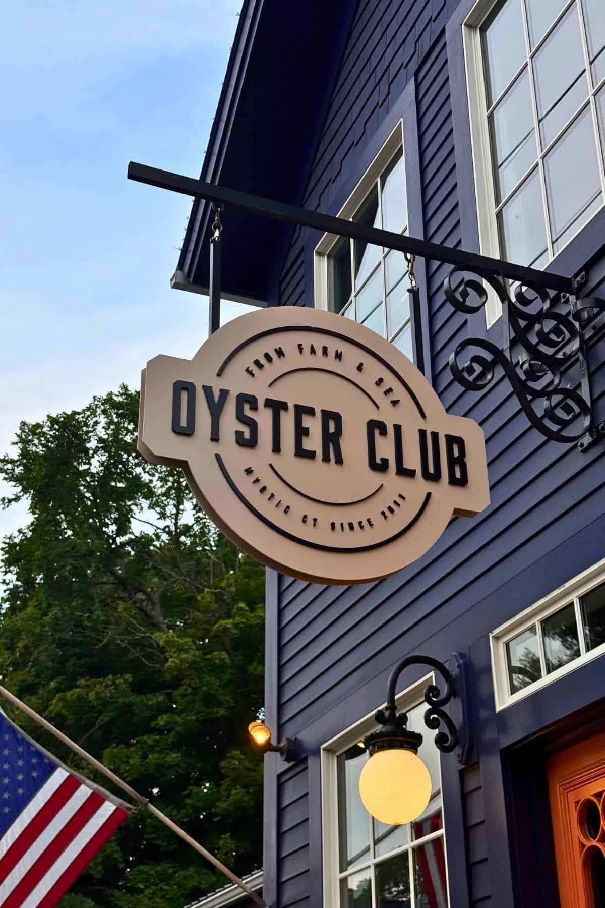 Round sign for Oyster Club restaurant in Mystic, CT, hanging from a blue building.