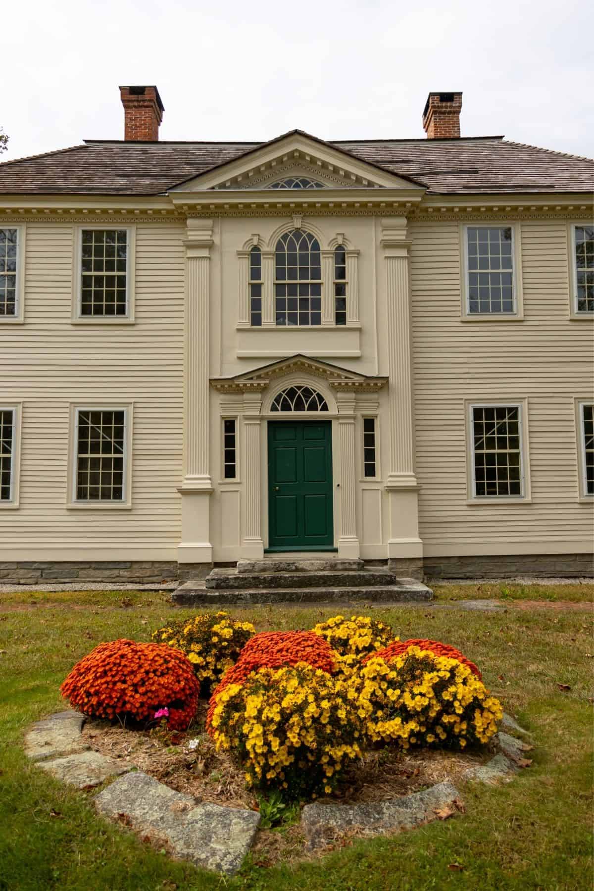 A two-story, cream-colored historic house with a central green door and a flower bed of red and yellow chrysanthemums in the foreground.
