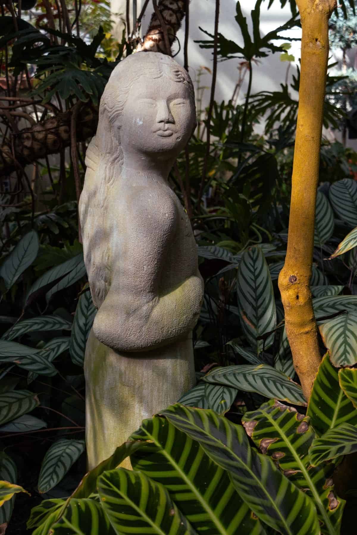 Stone statue of a young woman standing among tropical plants.
