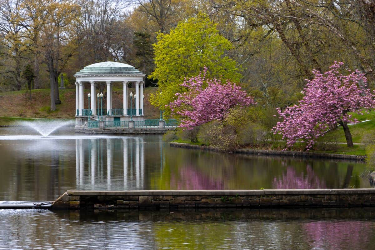 A domed pavilion stands beside a pond with a fountain and flowering trees in a park.