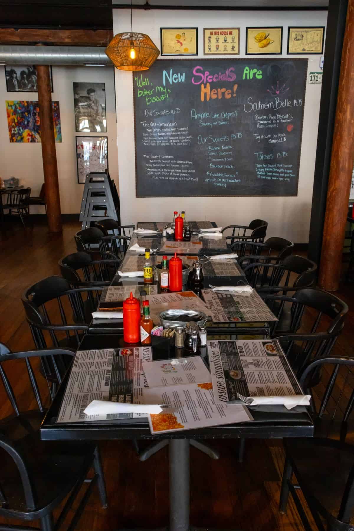 Dining room in a restaurant with tables set for guests, featuring a large chalkboard menu on the wall listing daily specials.