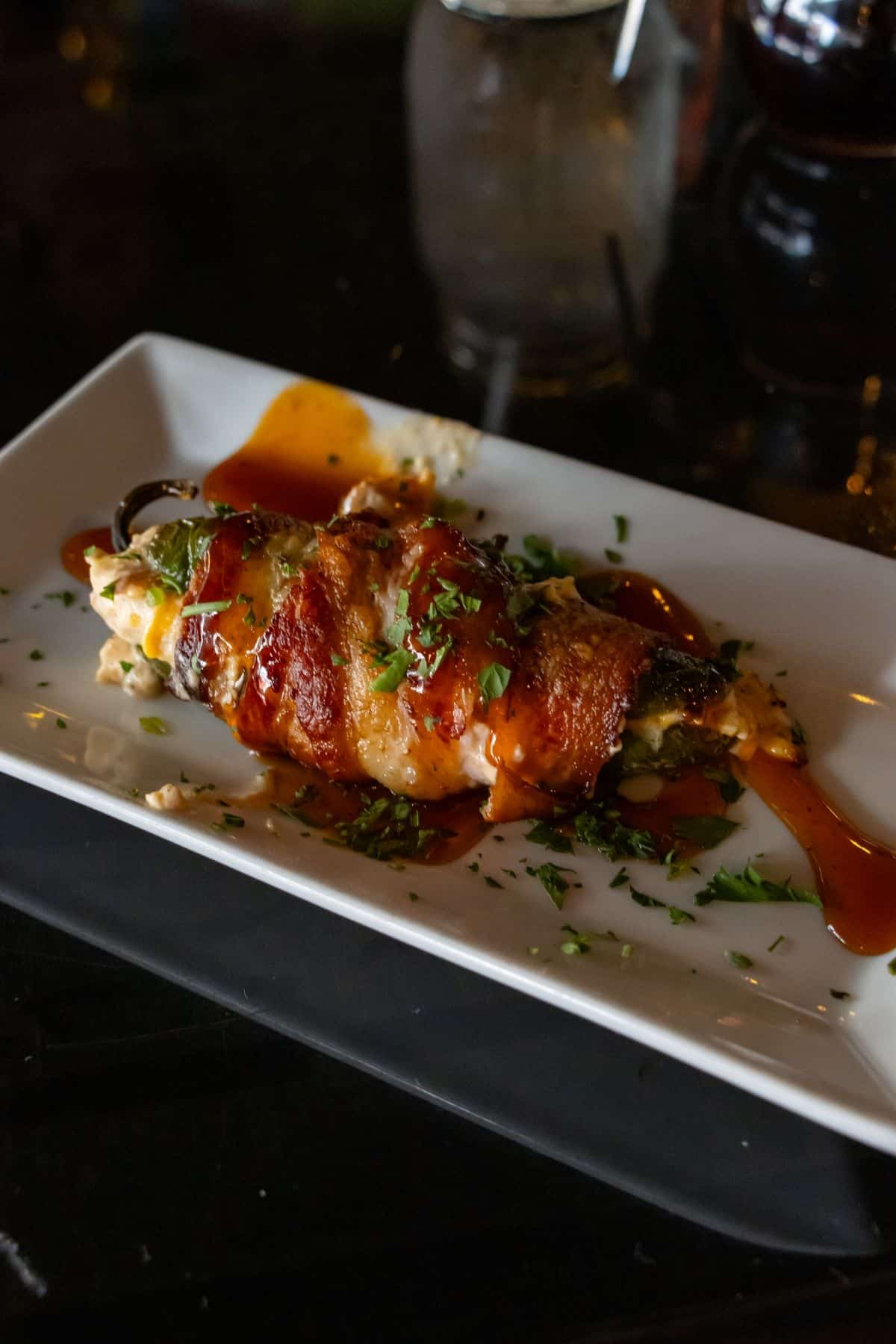 Bacon-wrapped jalapeño popper drizzled with sauce and garnished with herbs on a white rectangular plate.
